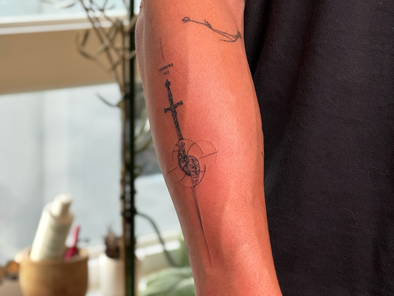 Fine-line sword piercing an anatomical heart inside a dotted ring, captioned Hebrews 4:12, on the forearm